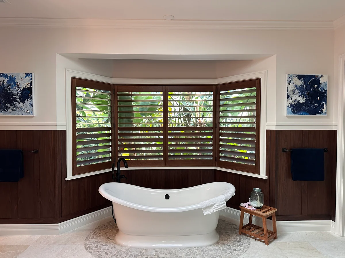 Stained wood plantation shutters around a bay window above a freestanding bathtub